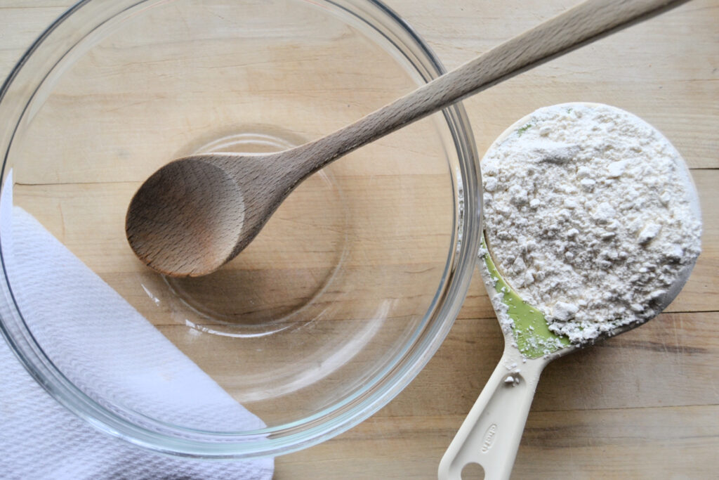 Empty glass bowl, wooden spoon, and measuring cup with flour inside