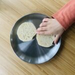 Child’s hand grabbing half a sourdough english muffin from a stainless steel plate