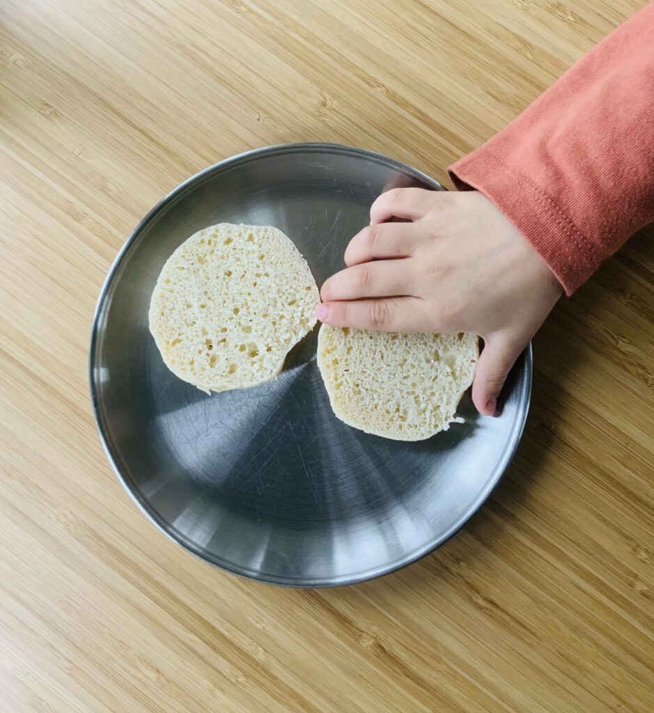 Child’s hand grabbing half a sourdough english muffin from a stainless steel plate